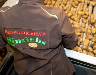 Eine Person in brauner Jacke steht vor einem Förderband voller Kartoffeln.