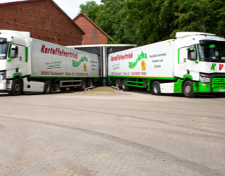 Zwei Lastwagen mit grüner und weißer Beschriftung stehen nebeneinander zur Beladung auf einem gepflasterten Platz vor einem roten Backsteingebäude und Bäumen im Hintergrund.