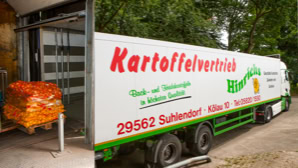 Ein LKW mit der Aufschrift "Kartoffelvertrieb Hinrichs" steht bereit zur Warenverladung, daneben sind gestapelte Säcke mit Kartoffeln auf einer Palette zu sehen.