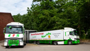 Zwei Lastwagen mit grüner und weißer Lackierung stehen nebeneinander auf einem Parkplatz, umgeben von Bäumen und einem Backsteingebäude.